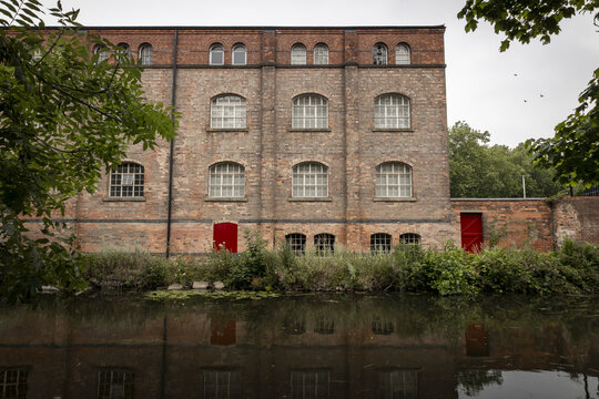 View of an old industrial brick building with arched windows and red doors situated along a calm canal in Nottingham, England, United Kingdom.