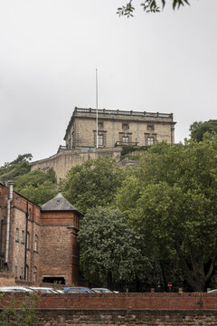View of the historic Nottingham Castle perched on a rocky hill surrounded by lush green trees and a brick wall in the foreground in Nottingham, England, United Kingdom.