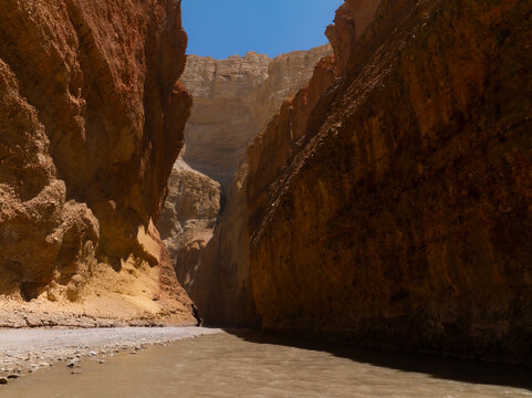 View of a deep canyon with towering orange-brown rock walls and a flowing river under a clear blue sky in Nepal.