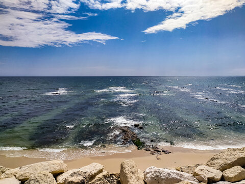 A serene, sunny beach showcasing clear blue ocean waves, sandy shore, and rocky foreground under a vibrant sky. Perfect for inspiring travel and mindfulness designs. No people.