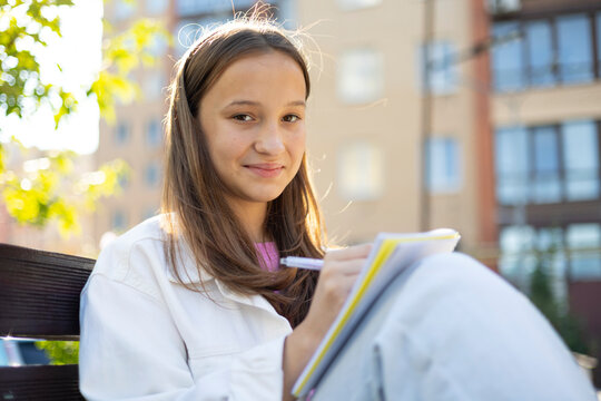 Smiling teenage girl making notes in a notebook while sitting outdoors in a casual urban setting. Education, studying, planning and lifestyle concept.