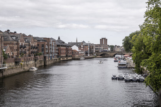View of the River Ouse in York, featuring historic brick buildings, a stone bridge, and small boats moored along the riverbank, York, England, United Kingdom.