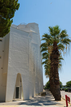 View of the distinctive white modern architecture with arched entrances and a row of tall palm trees under a clear blue sky in La Grande-Motte, Occitanie, France.