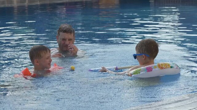 Energetic water play between father and two boys in clear blue pool. Outdoor activity, active childhood and memorable family vacation concept. Water blasters play in outdoor swimming pool