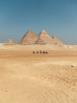 View of Giza Necropolis with camels and riders traversing the desert sands under a clear blue sky in Al Haram, Giza Governorate, Egypt.