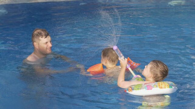 Two brothers laugh while playing with their father using colorful water blasters in clear blue pool. Family teamwork, active childhood and summer vacation concept. 