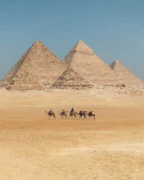 View of Giza Necropolis with people riding camels across the sandy desert landscape under a clear blue sky in Al Haram, Giza Governorate, Egypt.