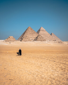 View of Giza Necropolis with a person in a black dress walking across the sandy desert under a clear blue sky in Al Haram, Giza Governorate, Egypt.