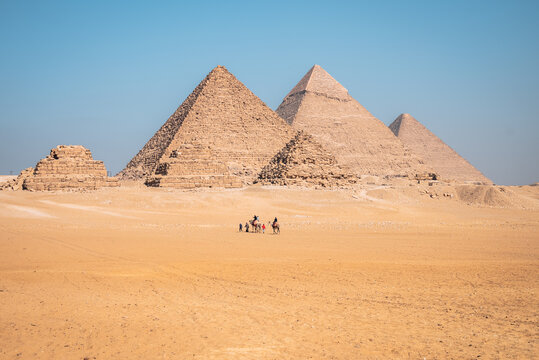 View of the Giza Necropolis with people riding camels across the sandy desert landscape under a clear blue sky in Al Haram, Giza Governorate, Egypt.