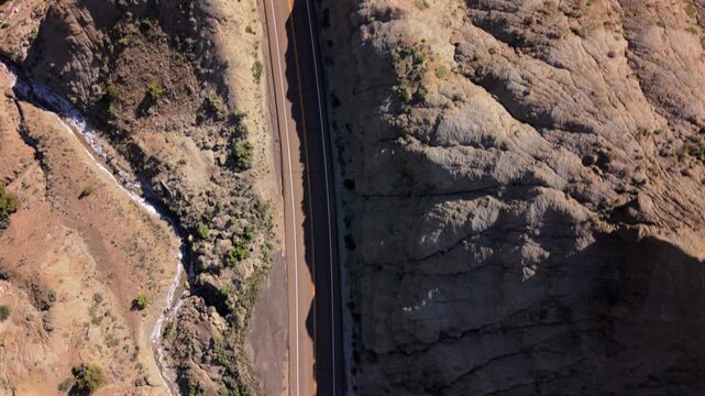 Aerial drone footage Utah Scenic Byway 12 reveals stunning desert canyon landscape with winding road cutting through rugged terrain. Empty highway stretches between rocky cliffs and dry creek bed.