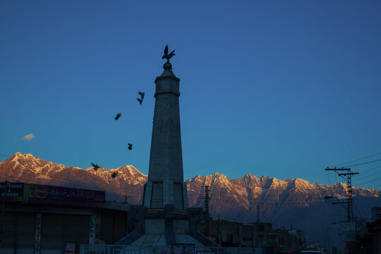 View of the Azadi Chowk monument with birds flying around the stone structure against a backdrop of snow-capped mountains under a clear blue sky in Skardu, Gilgit Baltistan, Pakistan.
