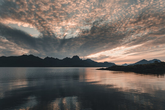 View of the calm waters of a fjord at sunset with silhouetted mountains under a dramatic textured sky in Holmstad, Nordland, Norway.