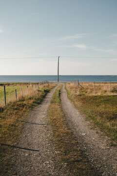 View of Andoya island with a dirt road leading towards the sea, flanked by grassy fields and a utility pole under a clear blue sky in Andoy, Nordland, Norway.