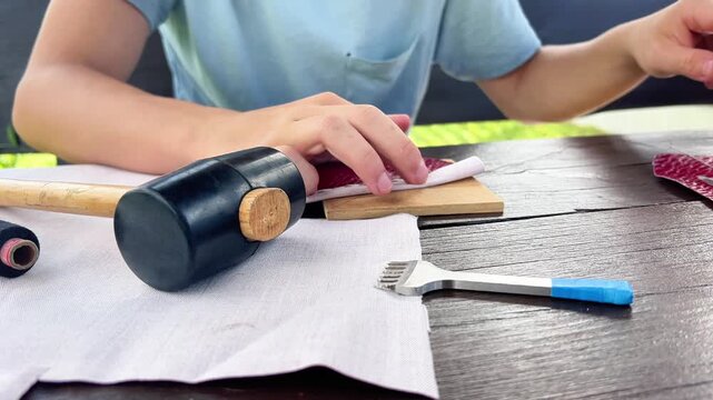 Close-up of artisan hands stitching genuine red leather using needle and black thread at a wooden table with leathercraft tools.