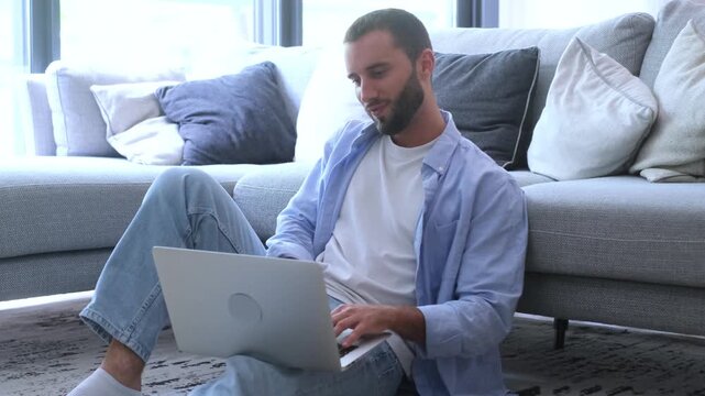 Casual young businessman working on a laptop computer while sitting on the floor in a modern living room, leaning against the sofa and smiling while enjoying his remote work lifestyle