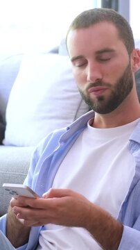 Focused handsome man sitting on the floor in his living room, texting messages, browsing social media, or shopping online using his smartphone with a concentrated expression on his face