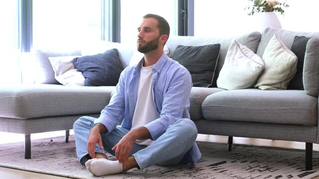 Calm young man practicing yoga meditation in the lotus position with eyes closed, sitting on the floor in his living room for mindfulness, relaxation, and stress relief