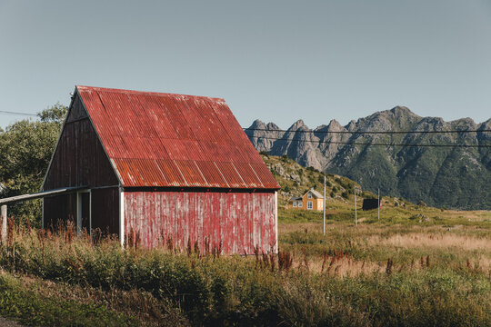 View of a rustic red barn with a corrugated metal roof set in a grassy field against a backdrop of rugged mountains in Hovden, Nordland, Norway.