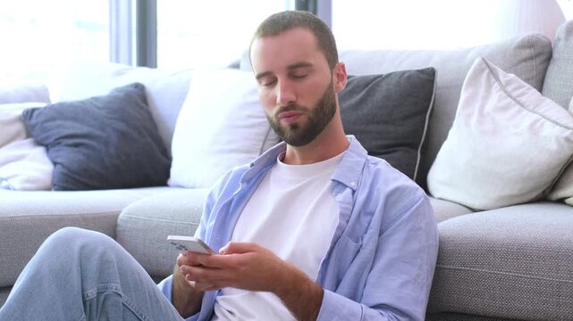 Focused young man with a beard sitting on the living room floor leaning against the sofa, scrolling and typing on his mobile phone while browsing social media or chatting online