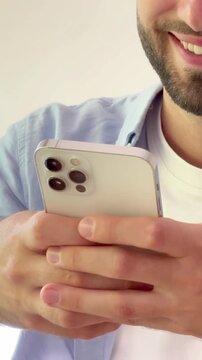Close-up of a smiling young man with a beard holding a modern smartphone, happily typing a message or browsing social media on a clean white background, showcasing digital communication