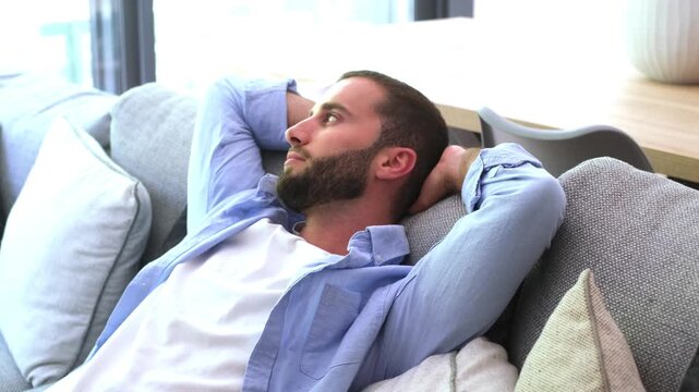 Young handsome man waking up from a nap on the couch in his living room, feeling rested and refreshed as he stretches his arms with a peaceful smile on his face