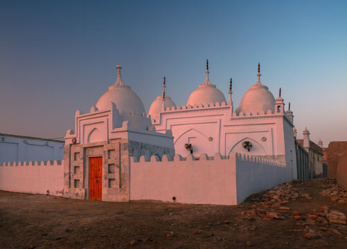View of the white mosque with multiple domes and a vibrant orange door illuminated by soft morning light in Ghaibi Dero, Sindh, Pakistan.