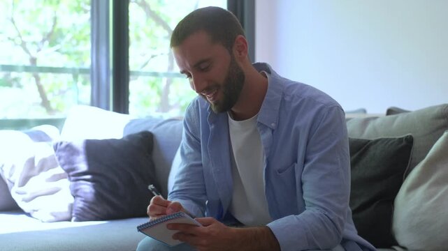 Young man sitting on a couch and writing in a notebook, smiling as he works on his creative ideas. Pensive male author or student planning and taking notes for a new project at home