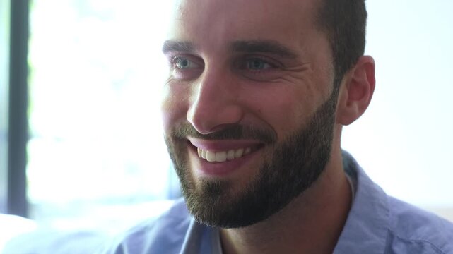 Close-up of a handsome young caucasian man with a beard and blue eyes, initially pensive, then looking away with a gentle smile, showing happiness and contentment in a brightly lit setting