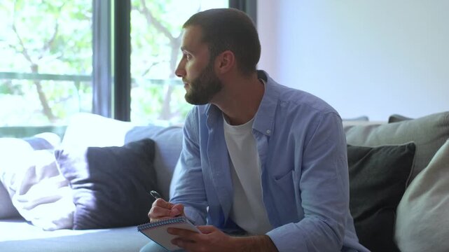 Pensive young bearded man sitting on a comfortable couch in his living room, writing his creative ideas, plans, or thoughts down in a notebook while looking for inspiration