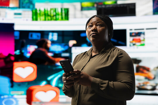 A woman browses a social media feed on a smartphone while standing before a video wall filled with multimedia content and online clips.