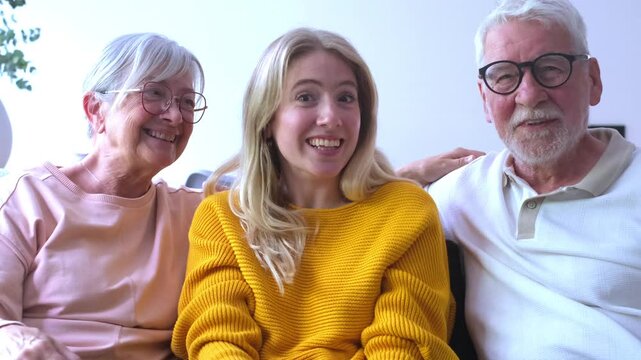 Smiling senior parents sitting on the couch with their young adult daughter while making a video call, waving and talking to the camera with cheerful expressions in their living room