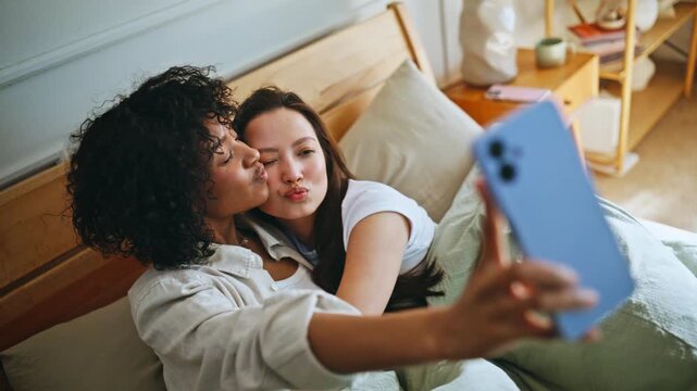 Lesbian couple taking selfie bed. Girlfriend kissing partner cheek morning.