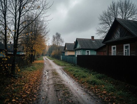 Empty rural road stretches through old village with wooden houses under grey cloudy sky. Autumn trees stand along dirt path covered by fallen yellow leaves. Weather gloomy yet peaceful in countryside