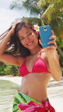 Girl taking phone selfie picture or video - Bikini travel woman on beach smiling happy using phone on Bora Bora. Vacation in Tahiti, French Polynesia. Vertical video of real woman without any makeup