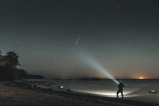 Merikula, Harju maakond, Estonia - 21 October 2025: View of a person standing on the beach at night, shining a flashlight towards the water under a starry sky with a visible comet.