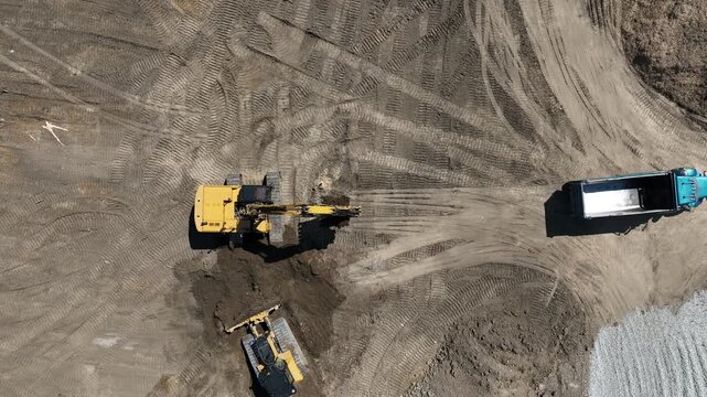 Aerial drone view of a hydraulic excavator loading soil into a dump truck while a bulldozer pushes earth at an active construction site during large-scale earthmoving operations.