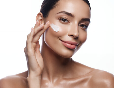 Beautiful young woman applying white moisturizing face cream to her cheek
