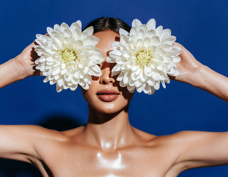 Woman Holding White Chrysanthemum Flowers Over Eyes Against Blue Wall