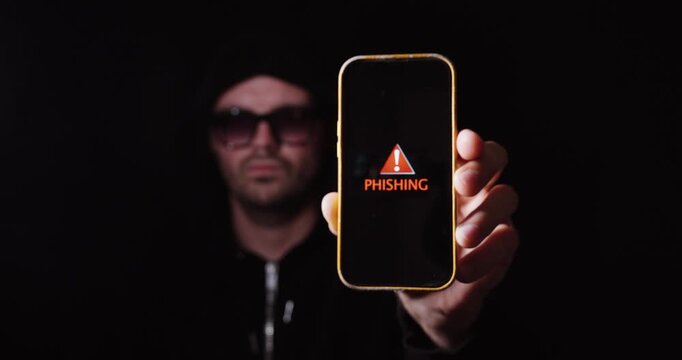A hooded cybercriminal holds a smartphone displaying a phishing warning message. Concept of phishing attacks, online fraud, cybersecurity, and digital threats.