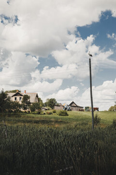 View of a white stork perched on a utility pole in a rural landscape with a farmhouse, a tractor, and green fields under a cloudy blue sky in Rezekne, Latvia.