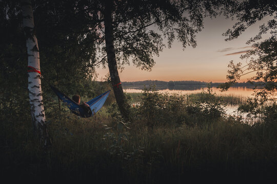 Zosna, Rezekne Municipality, Latvia - 15 July 2024: View of a person relaxing in a hammock strung between birch trees by the calm waters of Lake Zosna during a golden sunset.