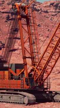 A large crane operates on a dry, rocky site. Workers are managing tasks while the machinery lifts heavy materials. The landscape shows red stones and clear skies.