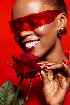 Close-up of a smiling black woman with dark skin wearing red translucent glasses and earrings holding a red rose. Concept of beauty, fashion, elegance, and self-expression in studio lighting.