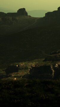 A view of a mountain landscape showing rocky formations and winding paths under a sunset sky. The shadows and light create patterns on the ground and rocks.