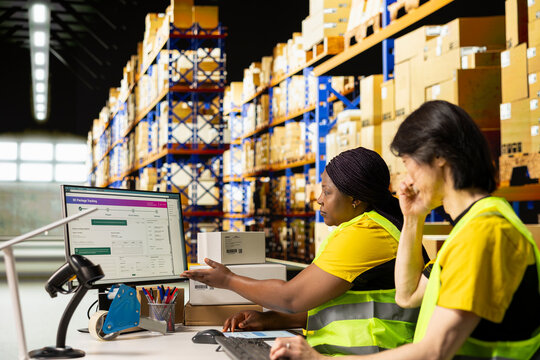 Multiethnic employees scan air waybill labels while a woman points to tracking software on screen, coordinating fast delivery among storage shelves.