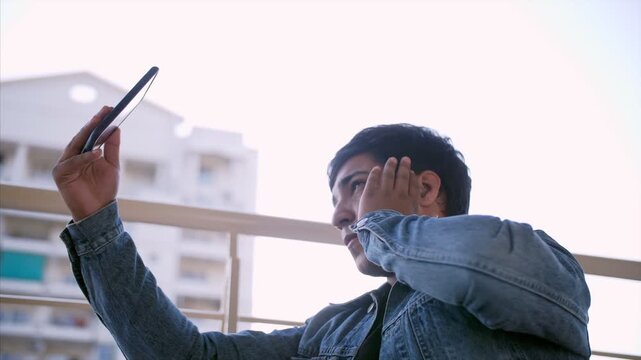 Young handsome man clicking pictures using his smartphone - technology concept. Side view shot of a carefree Indian guy taking selfies with his mobile phone while standing in his balcony - casual wear