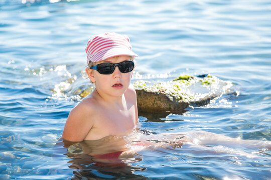 Young blond boy in pink striped cap and black sunglasses stands sadly in shallow blue sea near mossy rocksю