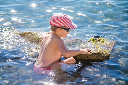 Young blond boy in striped cap and black sunglasses sits in clear blue sea water beside a mossy rock.
