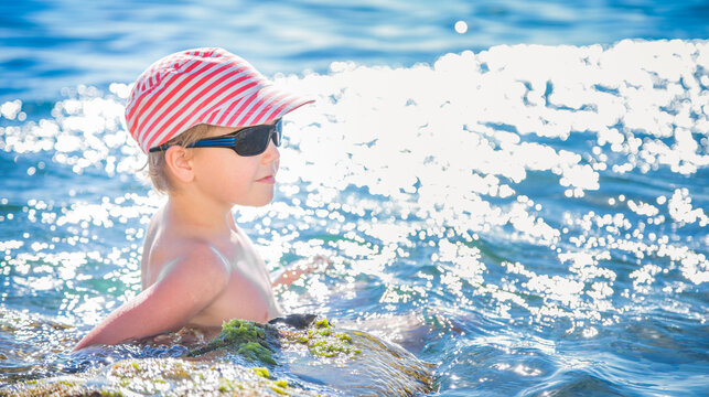 Young blond boy in striped cap and black sunglasses rests by a mossy rock in sparkling blue sea water.