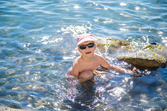 Young blond boy in striped cap and black sunglasses sits in clear blue sea water beside a mossy rock.